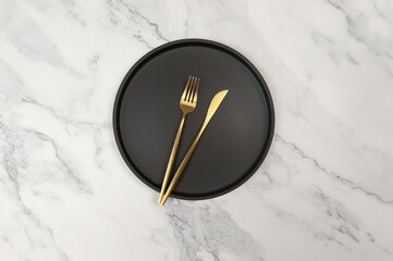 Top view of black plate and gold cutlery on white marble background. Table setting flat lay. Fork, knife and plate. Copy space.