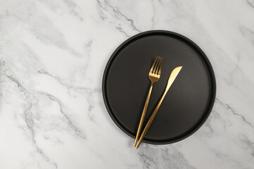 Top view of black plate and gold cutlery on white marble background. Table setting flat lay. Fork, knife and plate. Copy space.