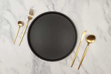Top view of black plate and gold cutlery on white marble background. Luxury table setting flat lay. Fork, knife, spoon, dessert spoon and plate. Copy space.