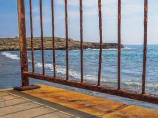 View from the pier through the rusty bridge fence over the Mediterranean Sea and rocks on a sunny day in Aye Napa, Cyprus.