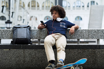 Skateboarder with headphones using mobile phone on bench with backpack. © juannovakosky
