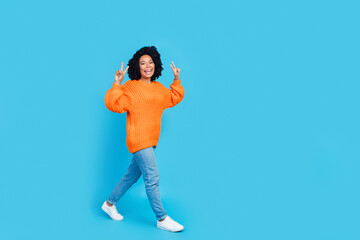 Fototapeta na wymiar Full size photo of attractive young woman walking show v-sign symbol wear trendy knitwear orange clothes isolated on blue color background