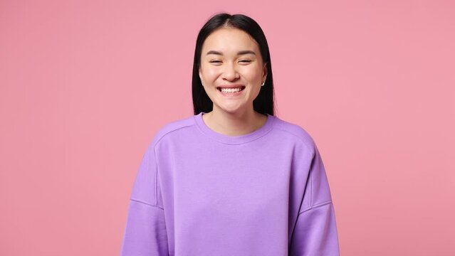 Young smiling happy cheerful fun cool woman of Asian ethnicity she wearing purple sweatshirt look camera isolated on plain pastel light pink color wall background studio portrait. Lifestyle concept