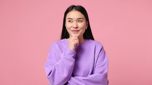 Young proactive puzzled woman of Asian ethnicity wear purple sweatshirt look around think scratches at temple comes up with idea raised finger get plan isolated on plain pastel light pink background
