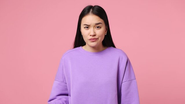 Young woman of Asian ethnicity wear purple sweatshirt showing finger gesture no I do not need it pointing thumb down dislike gesture isolated on plain pastel light pink background. Lifestyle concept