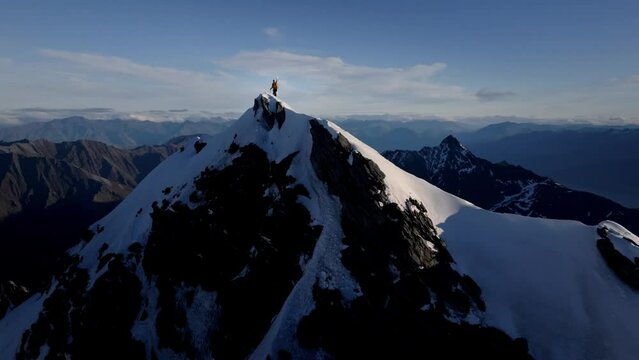 A majestic climber stands atop a Snowy Mountain in the wild reaches of nature with huge mountains and giant valleys in the background. Beautiful day at sunset. Drone shot.