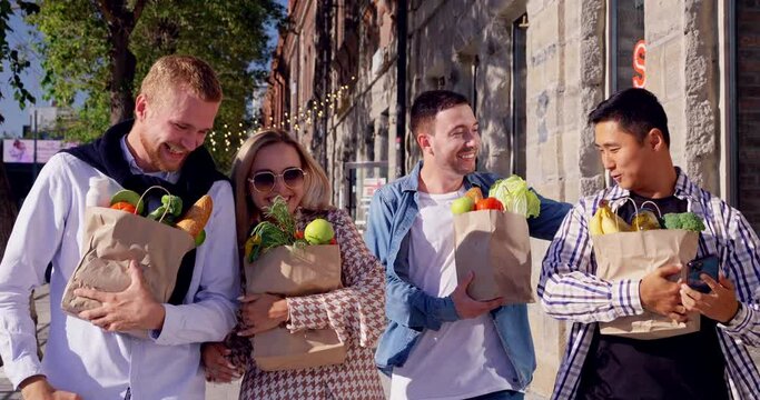 Group of four smiling friends together walk along street carrying bags with fresh food in sunny day. Shoppers happy with goods buys and groceries purchases