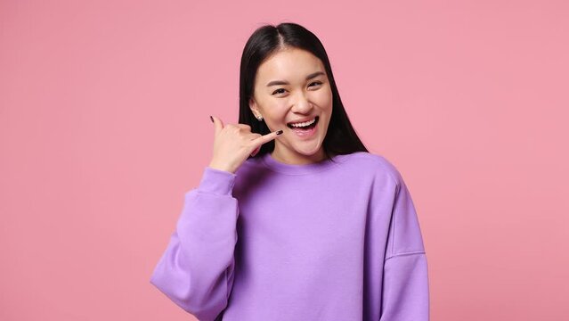 Young woman of Asian ethnicity wear purple sweatshirt point finger camera on you doing phone gesture like says call me back blinking isolated on plain pastel light pink background. Lifestyle concept