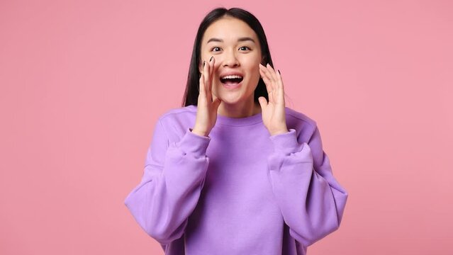 Excited happy fun cool young woman of Asian ethnicity wear purple sweatshirt scream hot news about sales discount with hands near mouth hurry up isolated on plain pastel light pink background studio