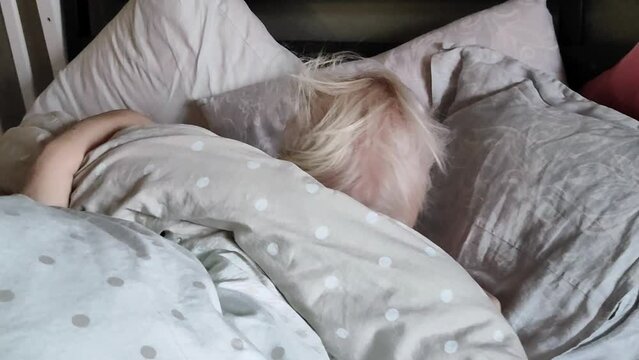 A little girl under two years old, sleepy in bed, crawls out from under the blanket and covers herself with the blanket again. Daytime sleep for babies.