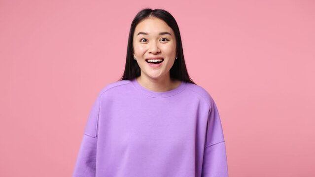 Young fun woman of Asian ethnicity wear purple sweatshirt look around for friend find waving meet greet with hand as notices someone say hello hi isolated on plain pastel light pink background studio