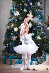 A little ballerina girl in a white tutu dress is holding her mother's finger and standing near the Christmas tree. 