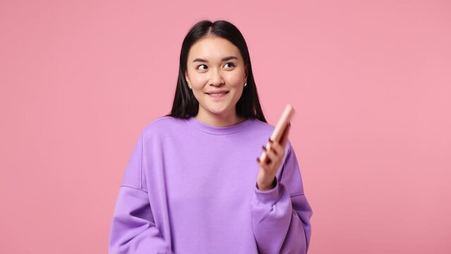 Happy young woman of Asian ethnicity wear purple sweatshirt hold use talk on mobile cell phone conducting pleasant conversation isolated on plain pastel light pink background studio. Lifestyle concept