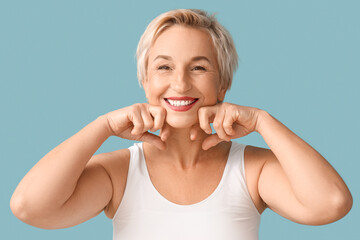 Fototapeta premium Beautiful mature woman doing face building exercise on blue background, closeup