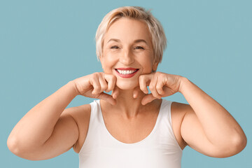 Beautiful mature woman doing face building exercise on blue background, closeup