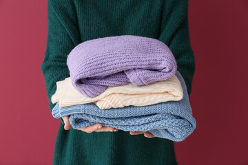 Woman holding stack of different stylish sweaters on burgundy background