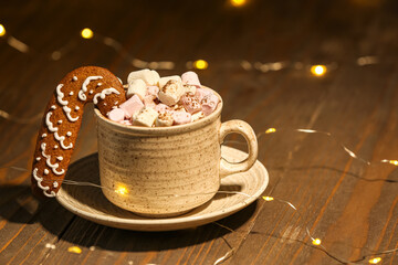 Cup of hot cocoa with gingerbread cookie and garland on wooden table against blurred Christmas background