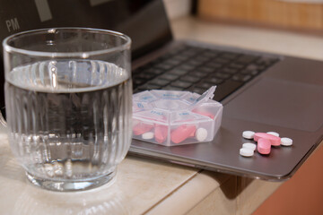Organizer weekly shots with glass of water on workplace background and laptop Closeup of medical pill box with doses of tablets for daily take medicine with white pink drugs and capsules. Medication