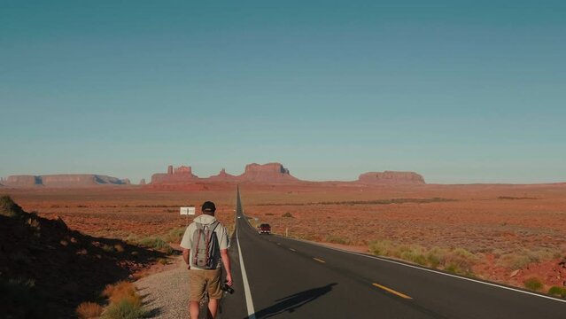 Camera follows hitch hiker man with backpack and camera walking along highway at sandstone desert of Arizona slow motion