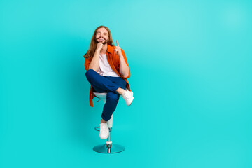 Fototapeta na wymiar Full length photo of cheerful man dressed orange shirt sit on chair indicating at discount empty space isolated on teal color background