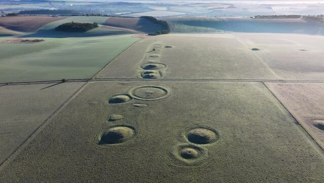 Wide high establishing aerial shot of burial mounds and barrows in low sun, Salisbury, UK