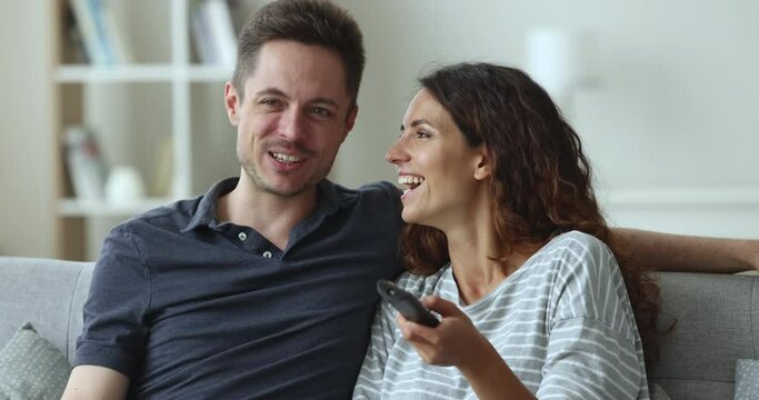 Happy attractive couple relaxing at home, watching sitcom series, funny movie on TV, discussing show, smiling, holding remote control, laughing, enjoying homey entertainment