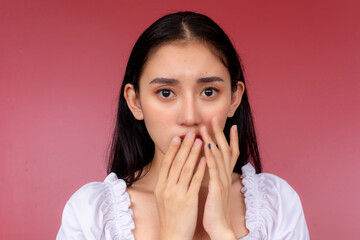 Portrait of a surprised and shocked Asian woman covering her mouth with hands against a vivid red...