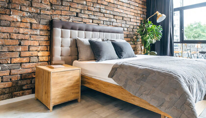 Wooden drawer nightstand near bed with grey fabric headboard. Loft interior design of modern bedroom with brown brick wall.