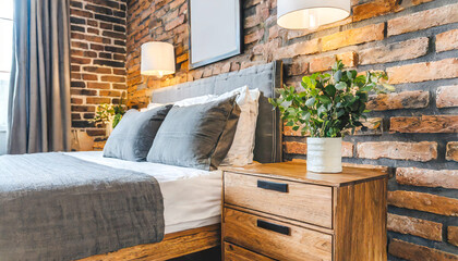 Wooden drawer nightstand near bed with grey fabric headboard. Loft interior design of modern bedroom with brown brick wall.