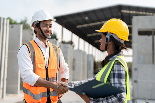Asian male and female civil engineers wearing vest and helmet safety handshake to work successfully at factory making precast concrete wall in construction site.