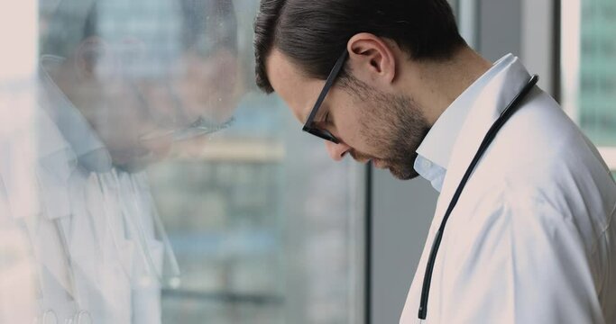 Depressed unhappy doctor man in white uniform and glasses standing at window, looking away, closing eyes, bending head down, thinking on medical mistake, problems, loss of patient, feeling despair