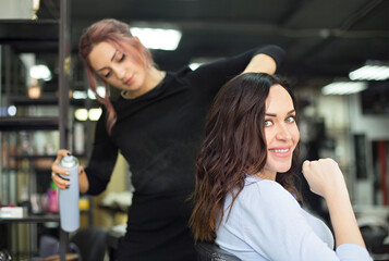 Young female hairdresser fixing hair of gorgeous woman with hairspray