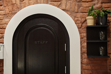 Wooden black half-round brown door with sign staff in brick wall.With shelf on wall with plant on it.