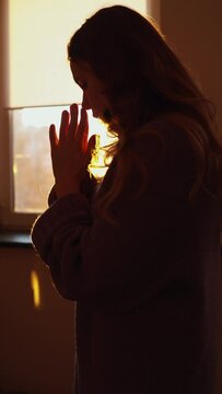 beautiful girl stands in front of the window at sunset and prays with folded hands. vertical video. meditation at home