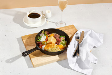 Sunny-side-up eggs in a skillet with roast beef, vegetables, and ciabatta, Americano coffee, and a glass of sparkling wine