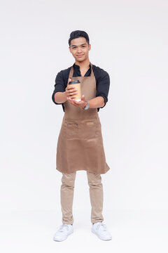 A handsome barista offering you a hot cup of coffee. Showing a lot of friendliness and hospitality. Wearing a brown apron. Full body studio shot, with white background.