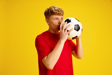 Sports betting. Young redhead guy with freckles in red t-shirt kissing soccer ball against yellow...