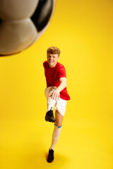 Dynamic image of young man in soccer sportswear, uniform training, hitting ball against yellow...