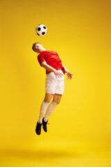 Fototapeta na wymiar Full-length image of young man in uniform, soccer player, enthusiast in motion, training, hitting ball with head over yellow studio background. Concept of active lifestyle, youth, hobby and emotions