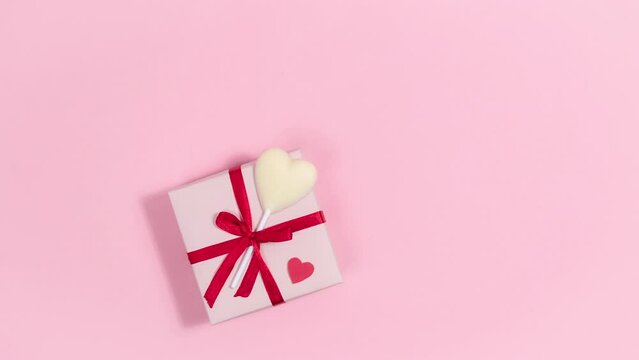 4k Gift box with red ribbon and with heart-shaped white chocolate candy. Near hearts fly by, symbols of love. Valentine's day greeting card. Pink background. Copy space. Stop motion animation.