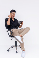 Excited young Filipino man sitting on an office chair, fist pumping while looking at his phone, portraying success and joy.