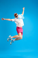 Obraz premium Full-length image of young happy, positive man in summer trip, jumping, having fun against blue studio background. Concept of emotions, youth, leisure time, summer vacation, travelling