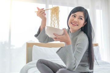 Beautiful asian woman eating chicken instant noodles soup in bed. Girl teenager watching sitting...