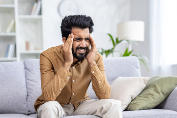Indian man experiencing stress and discomfort, touching his temples, sitting on a sofa in a living room apartment setting.