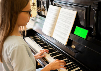 Young girl sitting at black piano using smartphone with green screen to studying playing piano.