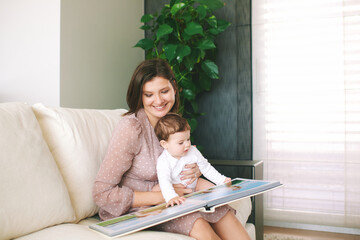 Portrait of young mother and adorable baby watching family album together, sitting on white sofa in living room