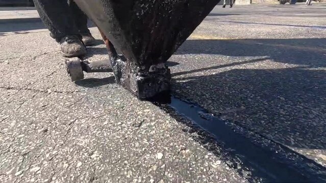 A worker repairs old asphalt. A worker pours bitumen into a crack in the asphalt.
