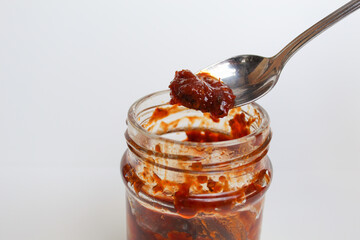 Sambal is an Indonesian chilli sauce or paste. On a silver spoon, taken from a transparent glass jar. Isolated on white background - obrazy, fototapety, plakaty
