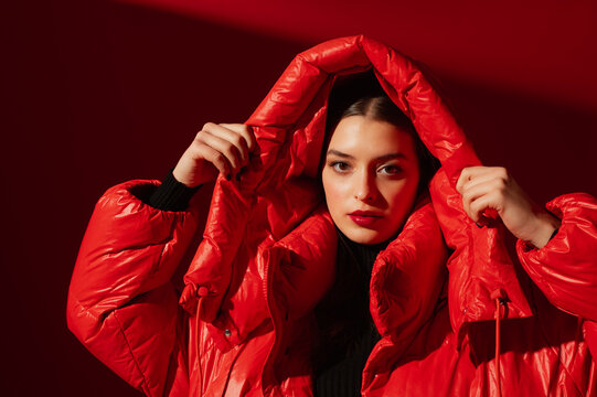 Fashionable confident woman wearing trendy oversized red puffer down jacket with hood,  posing on red background. Close up studio fashion portrait
