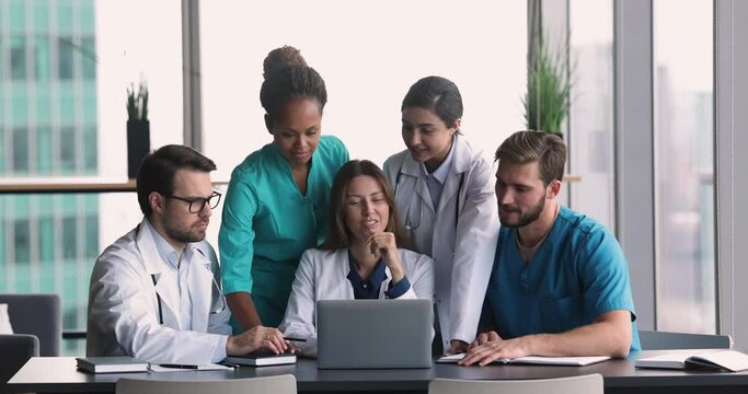 Diverse team of cheerful young doctors talking at laptop, watching online content, pointing at screen, speaking, smiling, laughing brainstorming on medical application for job